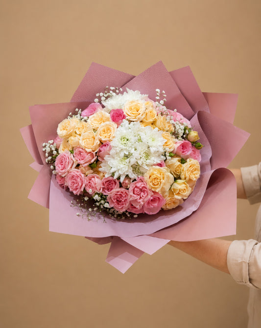 A large, vibrant bouquet of assorted flowers wrapped in pink paper, held by a person whose hands and arms are visible on the right side, set against a solid beige background.