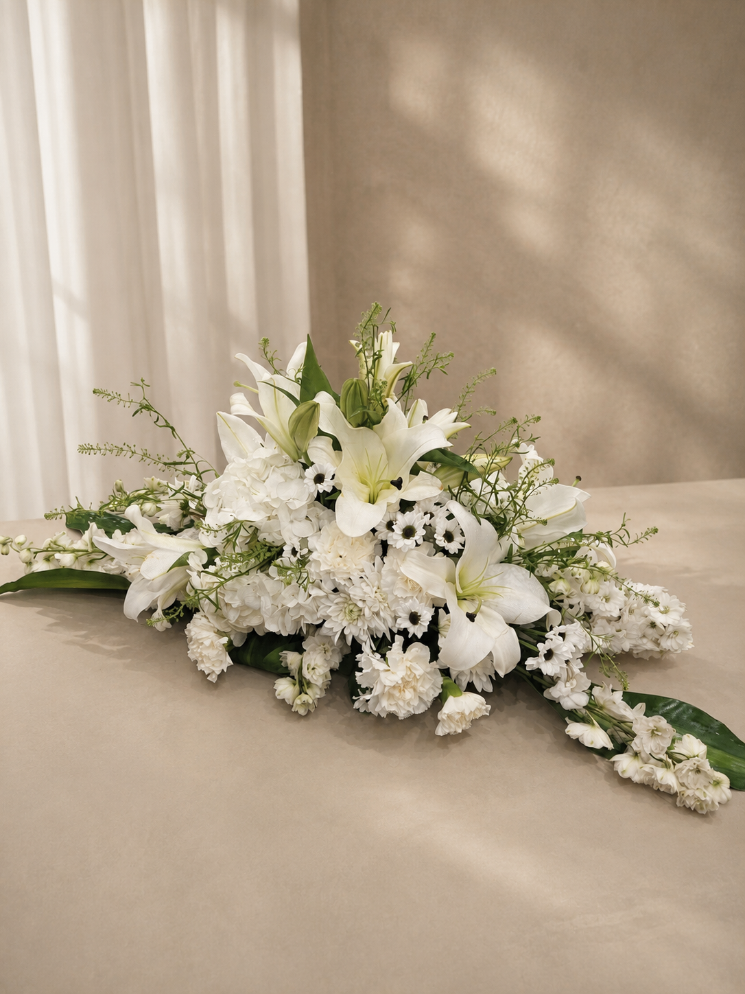 A white floral arrangement featuring lilies, hydrangeas, and green foliage, styled on a beige surface against a neutral beige background with soft window light shadows.
