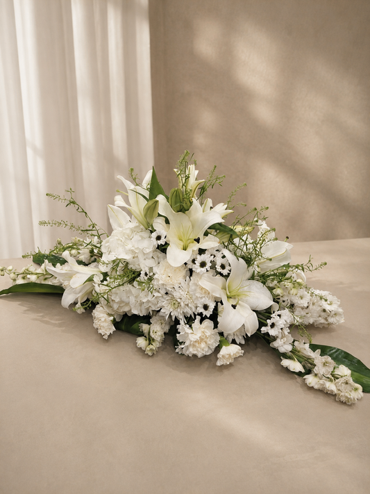 A white floral arrangement featuring lilies, hydrangeas, and green foliage, styled on a beige surface against a neutral beige background with soft window light shadows.